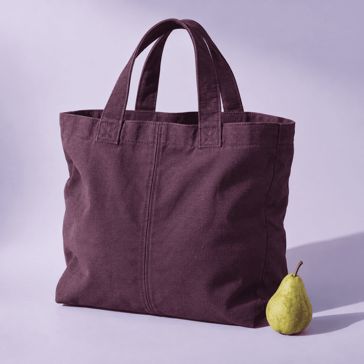 A deep plum cotton canvas Everyday Tote with wide handles beside a green pear